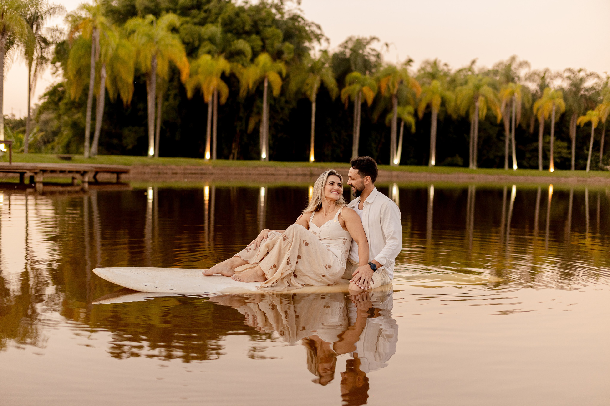 Casal sentado em prancha de SUP no meio do lago ao entardecer, com reflexo das palmeiras na água no Resort das Oliveiras