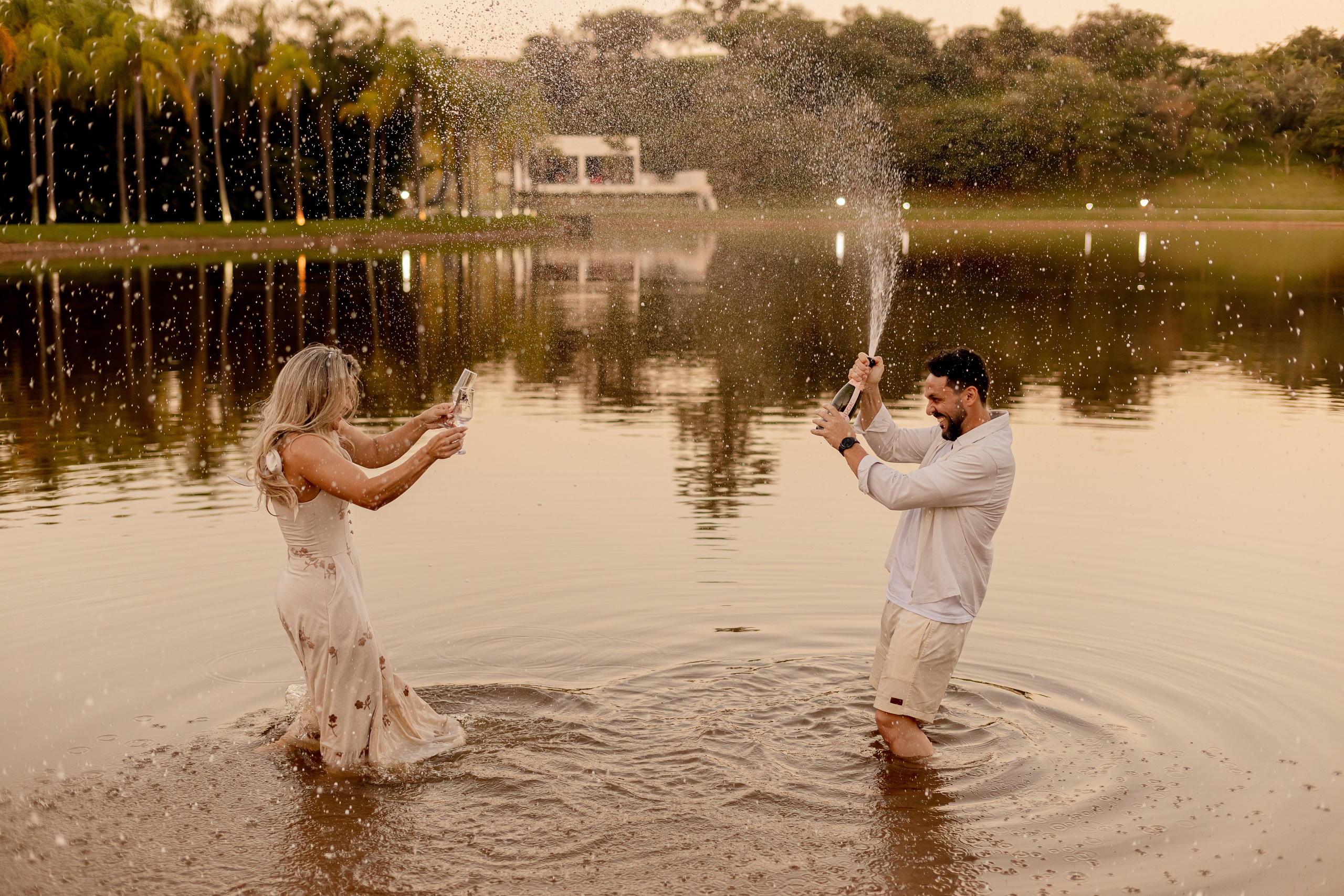 Casal celebrando com champanhe dentro do lago no Resort das Oliveiras ao entardecer, com espirros d'água e sorrisos