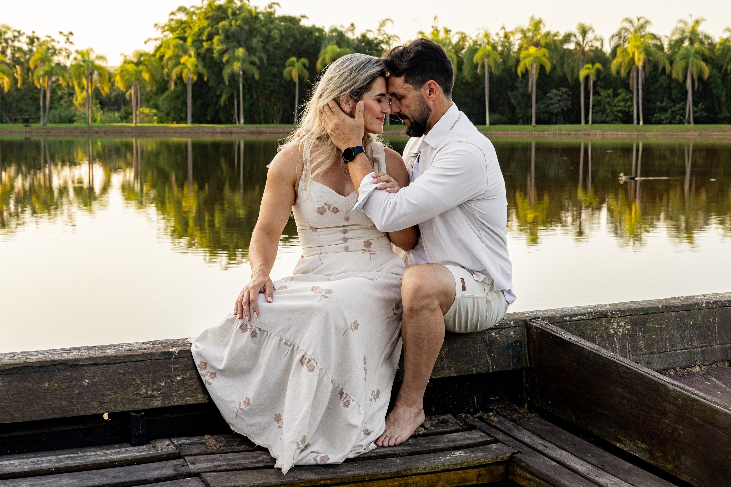 Casal sentado na borda de um barco de madeira se olhando com o pôr do sol dourado e o lago do Resort das Oliveiras ao fundo