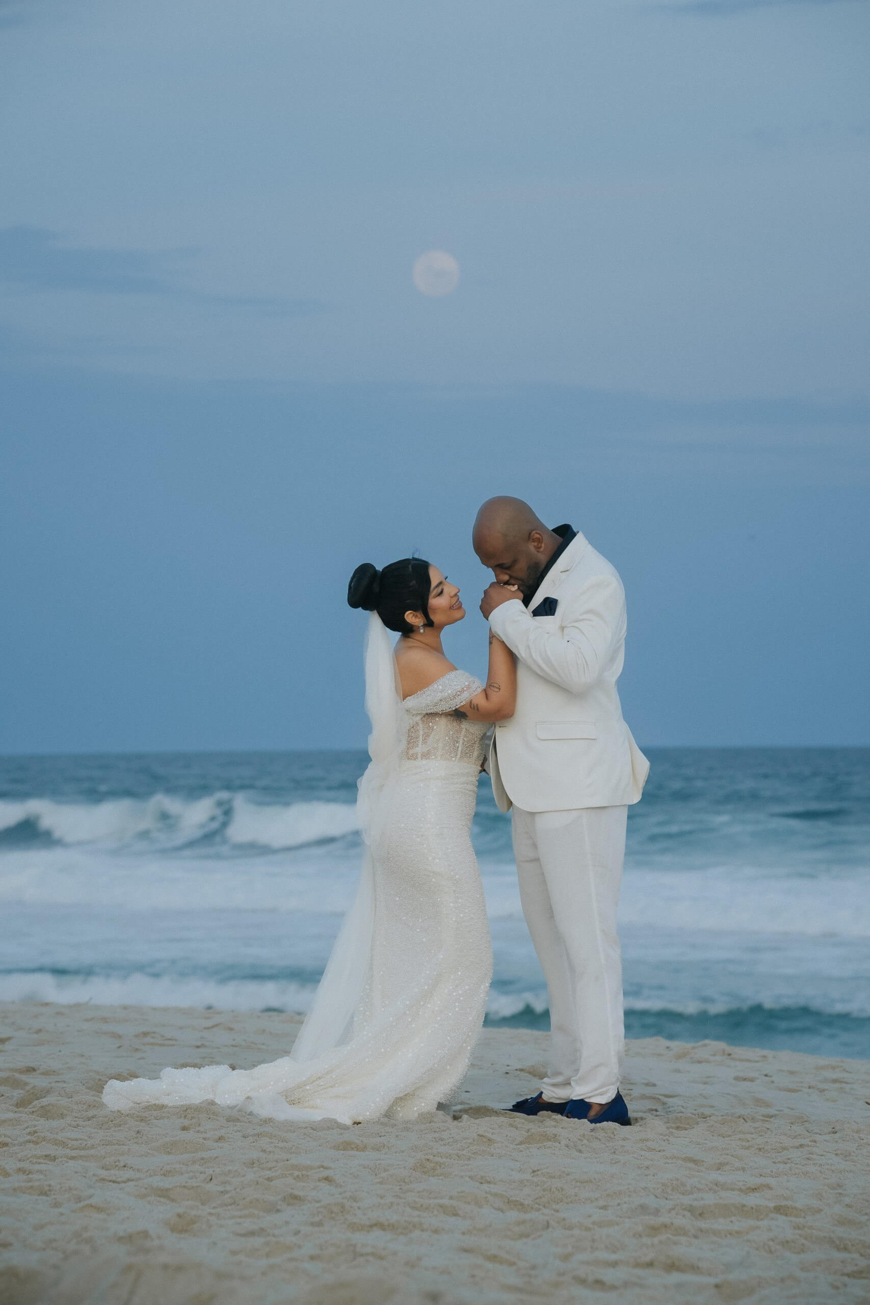 Noivo beijando as mãos da noiva em casamento na praia com a lua e o mar ao fundo